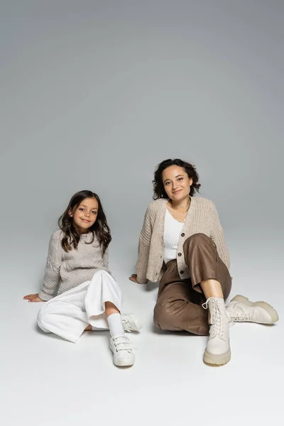 Brünette Mutter und Tochter in Strickwaren und Hosen auf grauem Hintergrund sitzend — Stockfoto