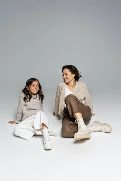 Smiling woman looking at trendy daughter while sitting on grey background — Stock Photo