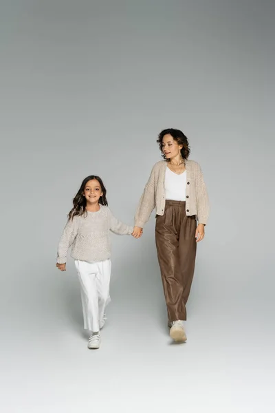 Full length of mom and daughter in warm knitwear and pants walking while holding hands on grey background — Stock Photo