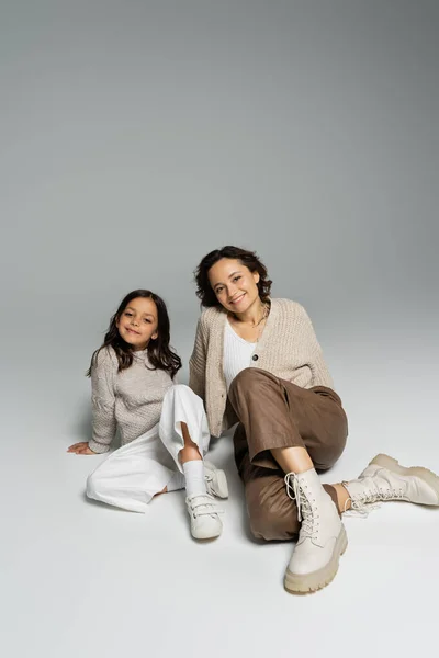 Comprimento total de mãe e filha em malhas e calças sentadas em fundo cinza — Fotografia de Stock