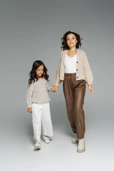 Donna alla moda sorridente alla fotocamera mentre si tiene per mano con la figlia e cammina su sfondo grigio — Foto stock