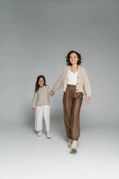 Glückliche und stilvolle Frau, die in die Kamera schaut, während sie ihre Tochter an der Hand hält und auf grauem Hintergrund geht — Stockfoto