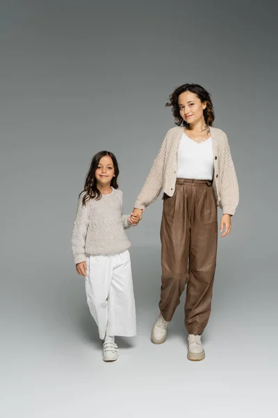 Mère avec enfant en tenue d'automne élégante tenant la main et regardant la caméra tout en marchant sur fond gris — Photo de stock