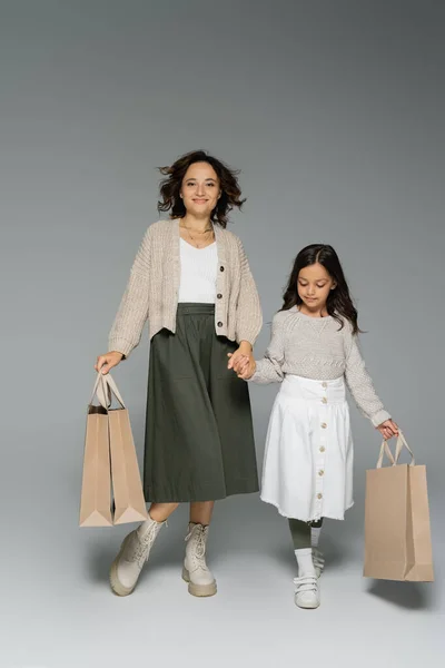 Longitud completa de la mamá y la hija en faldas cogidas de la mano mientras lleva bolsas de compras sobre fondo gris - foto de stock