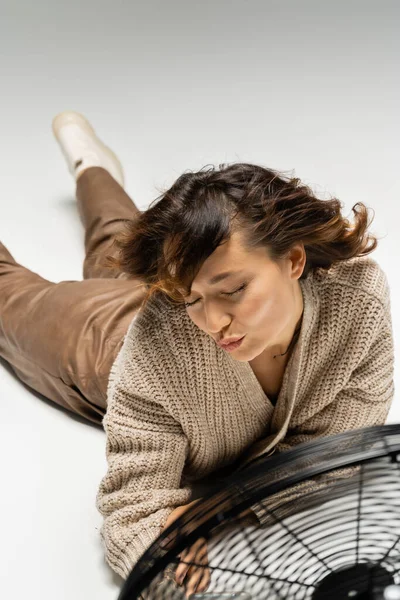 Женщина в теплом кардигане надувает губы, лежа рядом с дующим электрическим вентилятором на сером фоне — стоковое фото
