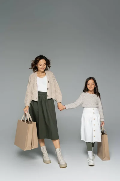 Longitud completa de la mujer y el niño en prendas de punto y faldas tomados de la mano y caminando con bolsas de compras sobre fondo gris - foto de stock