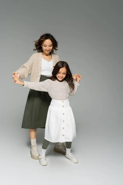 Happy mother and child in stylish clothes holding hands and dancing on grey background — Stock Photo