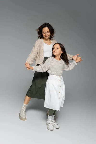 Happy girl and woman in trendy clothes dancing while holding hands on grey background — Stock Photo