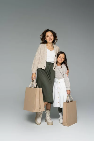 Mujer feliz en ropa de otoño elegante sosteniendo bolsas de compras y abrazando a la hija sobre fondo gris - foto de stock
