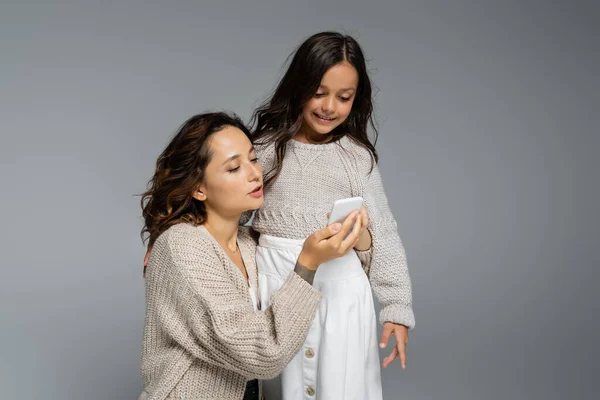 Brunette woman showing mobile phone to smiling daughter isolated on grey — Stock Photo