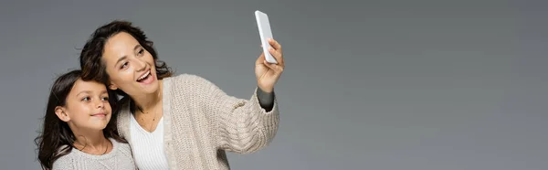 Mujer elegante y alegre tomando selfie con hija sonriente aislada en gris, bandera - foto de stock