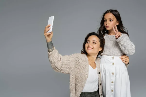 Trendy girl showing victory sign near smiling mom taking selfie on cellphone isolated on grey — Stock Photo