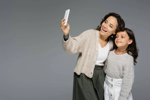 Donna eccitata scattare selfie su smartphone con figlia in abiti alla moda isolati sul grigio — Foto stock