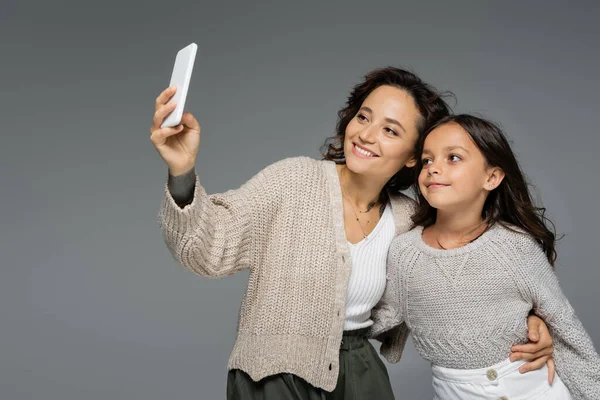 Happy mother and daughter in warm knitted clothes taking selfie on smartphone isolated on grey — Stock Photo