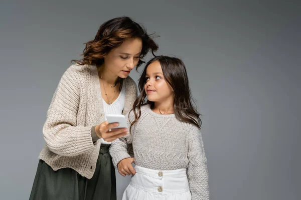 Улыбающаяся девушка в стильной осенней одежде смотрит на маму со смартфоном, изолированным на сером — стоковое фото