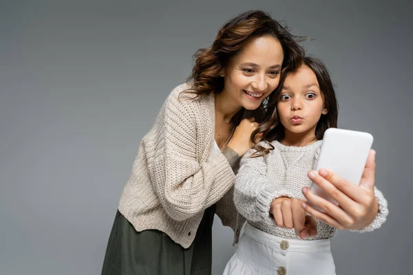 Amazed girl pointing at mobile phone near smiling mother in knitwear isolated on grey — Stock Photo