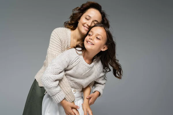 Happy woman with closed eyes hugging daughter in warm knitwear isolated on grey — Stock Photo