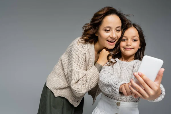 Chica sonriente mostrando teléfono móvil borroso a mamá sorprendida aislada en gris - foto de stock