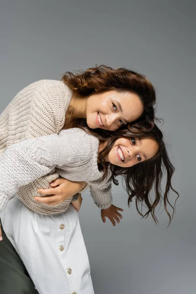 Cheerful woman embracing girl in trendy autumn outfit and smiling at camera isolated on grey — Stock Photo