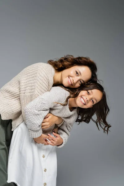 Cheerful woman and child in warm outfit embracing and smiling at camera isolated on grey — Stock Photo