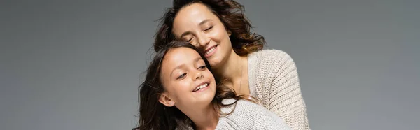 Mulher na moda sorrindo com os olhos fechados perto da filha feliz isolado em cinza, banner — Fotografia de Stock