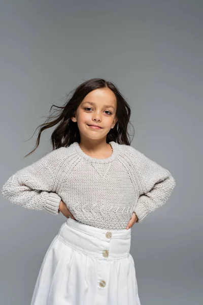 Trendy girl in knitted sweater and white skirt posing with hands on hips isolated on grey — Stock Photo