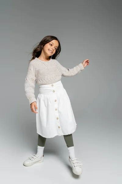 Pleine longueur de fille heureuse en pull chaud et jupe blanche posant sur fond gris — Photo de stock