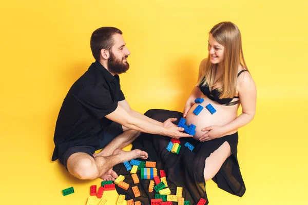 Marido e mulher jogar criança construtor. O cara faz um rosto sorridente do construtor na barriga de uma menina grávida. — Fotografia de Stock