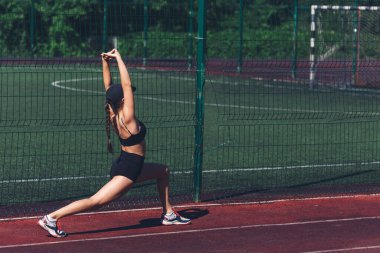 Genç bir kadın yaz aylarında kollarını spor sahasına uzatarak derin hamleler yapıyor.