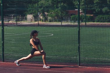 Genç bir kadın yaz aylarında spor sahasında derin hamleler yapıyor.
