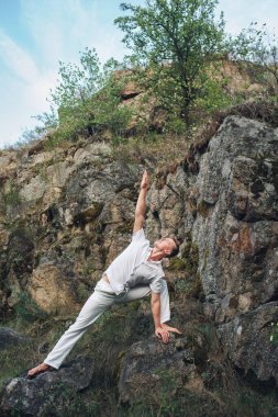 Doğada yoga yapan genç adam, kayaların yanında utthita trikonasana pozu veriyor.