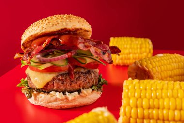 Kıpkırmızı arka planda salatalık, salatalık, domates, soğan ve pastırma ile dolu kalın, taze bir çizburger. Mısır koçanına süslenmiş.