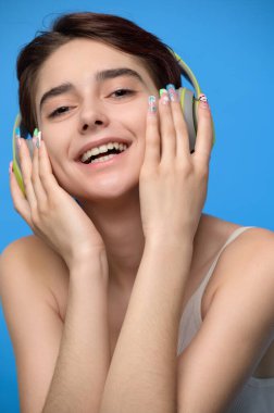 Portrait of young cute brunette with extravagant nail art listening to music using bluetooth wireless headphones, studio shot