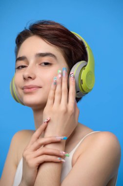 Portrait of cute young bare shoulders brunette with extravagant nail art listening to music using bluetooth wireless headphones, studio shot