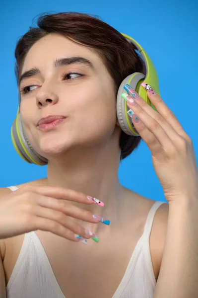 Portrait of cute young bare shoulders brunette with extravagant nail art listening to music using bluetooth wireless headphones, studio shot