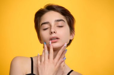 Romantic studio portrait of young pretty girl pouching her lips, short haircut and extravagant nail art, eyes closed, mouth slightly open, yellow colored background