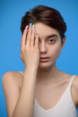 Skinny calm teenage girl cute girl hiding right half of her face with hand, at blue background