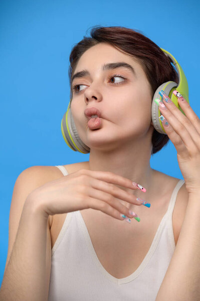 Милая юная брюнетка с короткой стрижкой делает рыбьи губы гримасой, слушая музыку с помощью беспроводных наушников