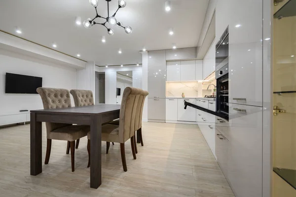 White spacious kitchen interior with dining room table and chairs, a television on the wall in the background and an oven with door open