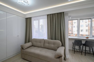 Kanepesi olan küçük bir stüdyo dairesi ve pencere kenarında sandalyeleri olan bir yemek tezgahı.