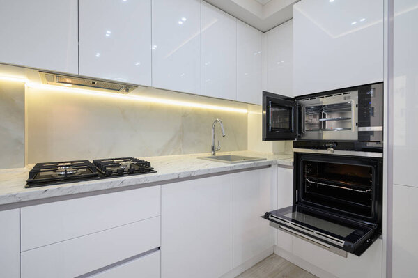 White modern kitchen with a stove, oven and microwave with open doors, glass electric kettle at worktop.