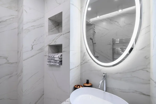 White bathroom interior with marble tiles on the walls and a round mirror