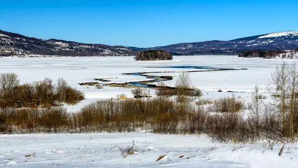 Arka planda tepeler ve göl adaları olan donmuş göle nehrin aktığı bir kış manzarası. Soğuk bir kış gününde Donmuş Göl.