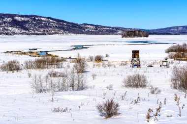 Kış manzarası, arka planında tepeler ve göl adası olan donmuş göle giren bir gözetleme direği ve akarsu akarsudur. Soğuk bir kış gününde Donmuş Göl.