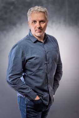 Sakallı, orta yaşlı bir adamın portresi özenle kameraya bakıyor. Gri renkli arka planda fotokopi odası var.