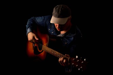 Kot pantolonlu bir gitarist siyah arka planda akustik gitar çekimi yapar. Gösterişsiz bir portre.