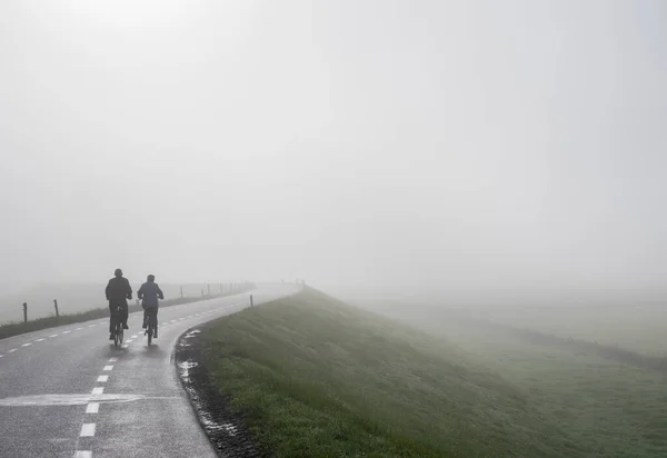 elderly couple on bike rides in mist on dike near river rhine in the netherlands on foggy autumn morning