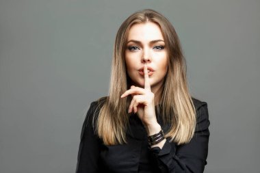 Genç bir kadın sessizlik işareti gösteriyor. Siyah gömlekli çarpıcı bir sarışın parmağını dudaklarına koydu. Sessizlik ve gizlilik. Gri arkaplan.
