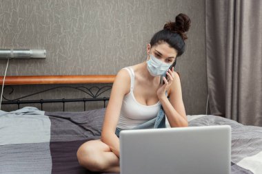 Tıbbi maskeli, telefonu ve laptopu olan genç bir kadın yatağın üzerinde oturuyor. Uzak çalışma ve kilitleme.