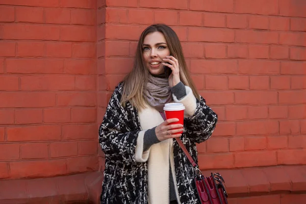 Young smiling woman with phone and coffee on a city street. Beautiful blonde in a warm stylish coat against a background of a red brick wall. Blogging and remote work.